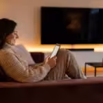 A woman relaxes on a sofa using a tablet in a cozy, modern living room with warm lighting.