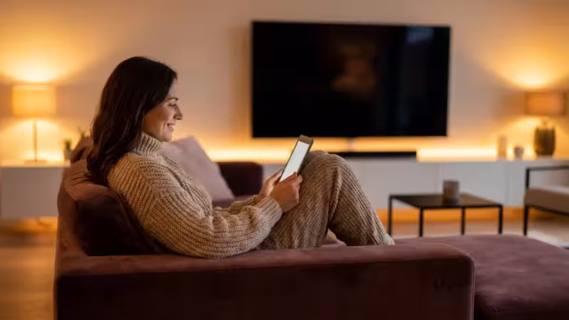 A woman relaxes on a sofa using a tablet in a cozy, modern living room with warm lighting.