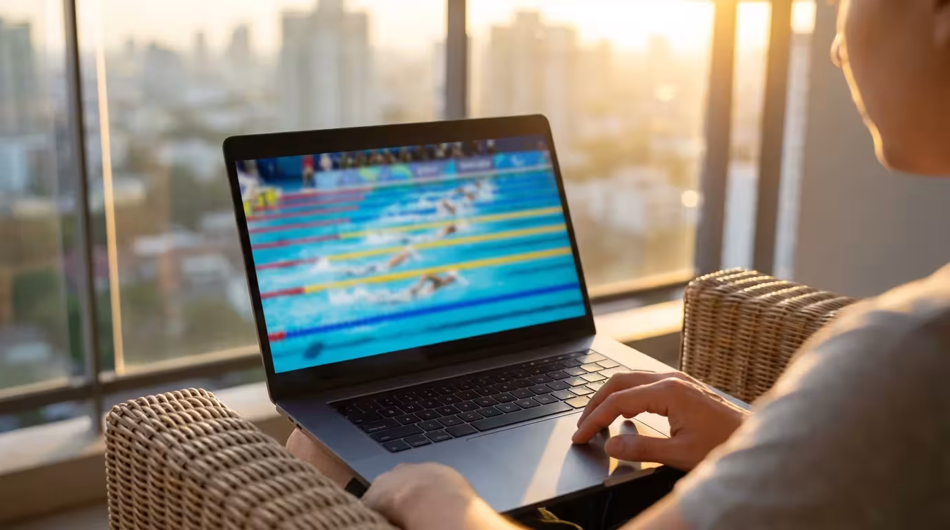 Over-the-shoulder view of a person watching sports on a laptop on a balcony during sunset.